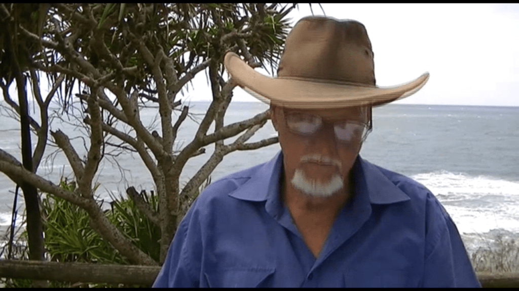 An older man wearing a large hat and glasses stands outdoors by the ocean, with trees in the background.